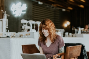 A redheaded woman in a cafe looking at her laptop and smiling.